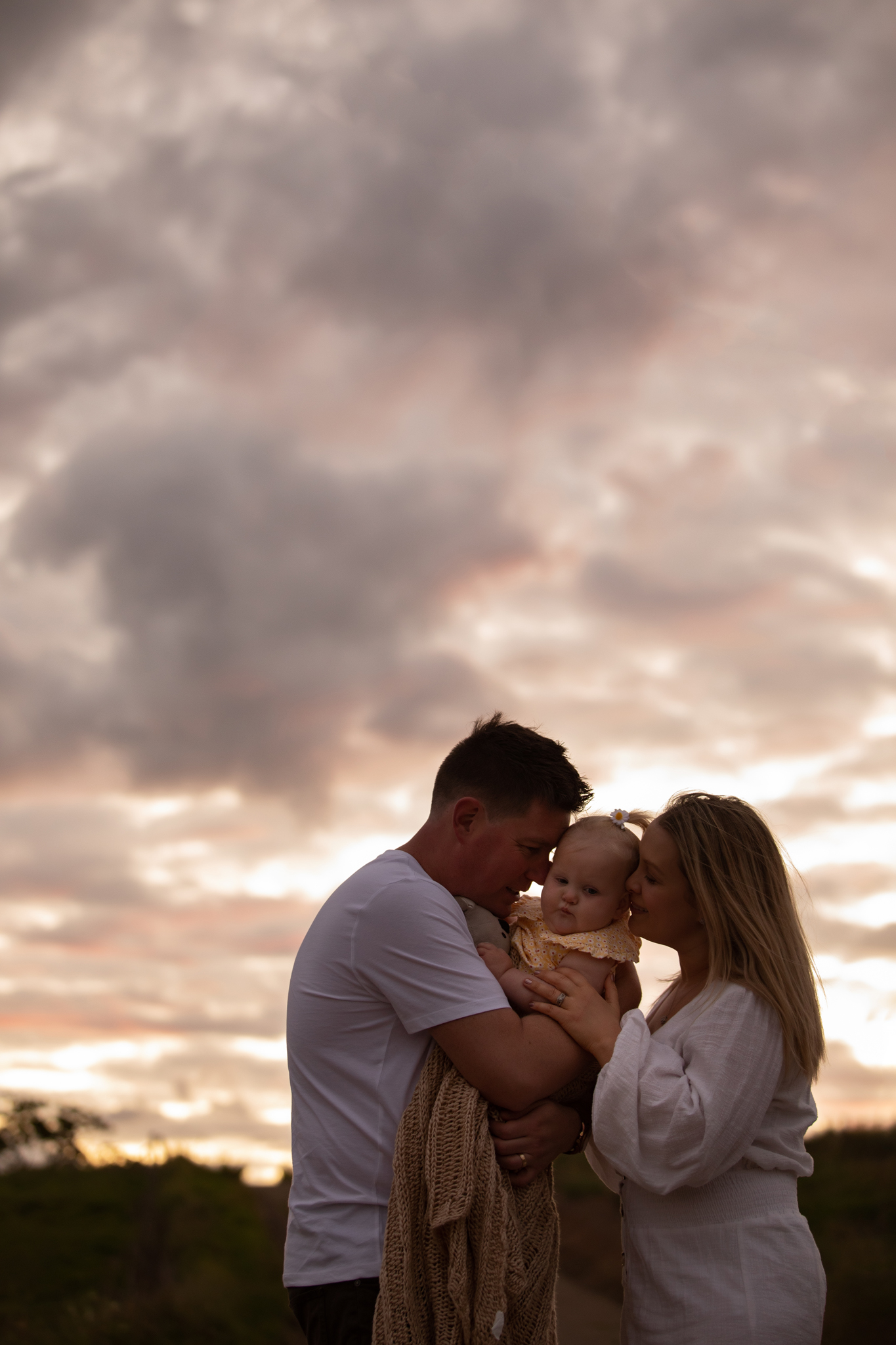 couple holding their baby close and hugging her, after sunset with cloudy skies